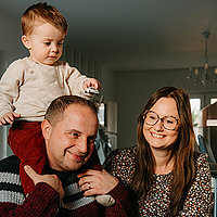 Theo mit seinen Eltern Familie: ein Mann hat ein kleines Kind auf den Schultern, daneben sitzt eine Frau in einem Blümchenkleid.