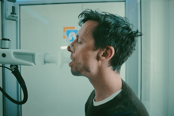 Mann mit dunklen kurzen Haaren sitzt in einem Glaskasten und hat ein Rohr im Mund, er scheint herein zu blasen. So läuft ein Lungenfunktionstest ab. 